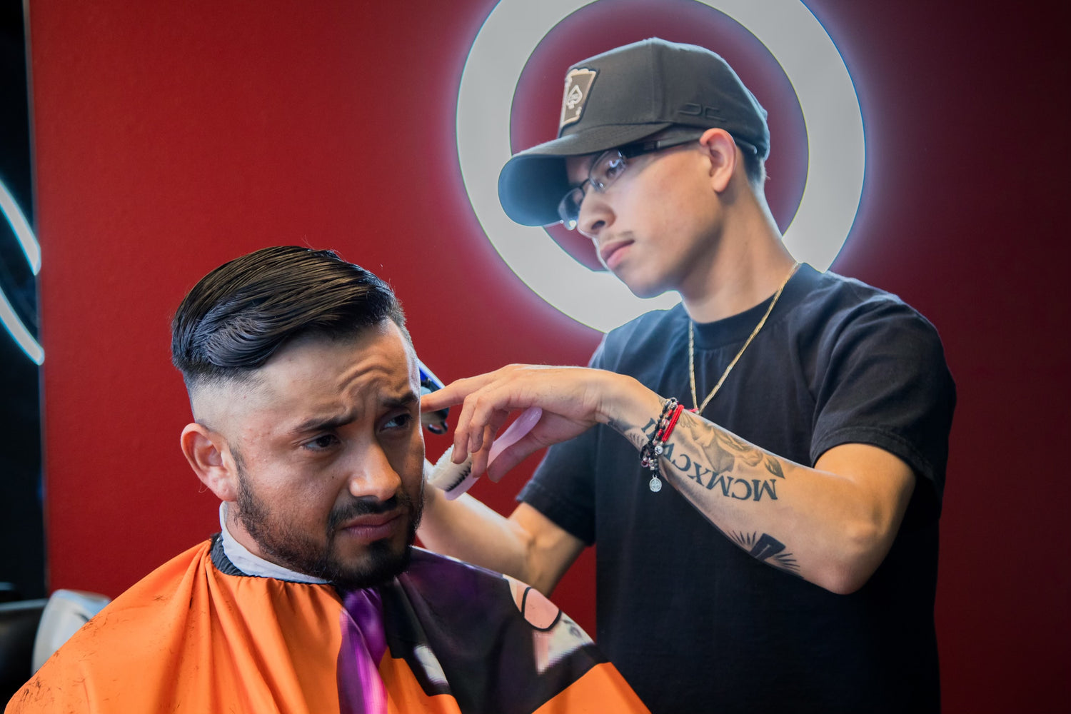 Barber with a black shirt and hat giving a client with an orange apron a haircut 