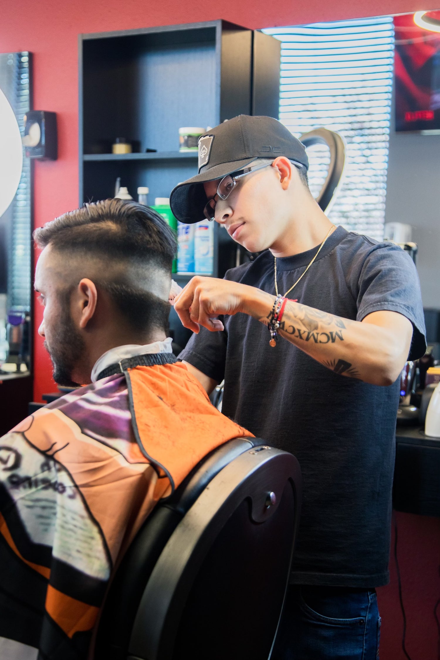 Barber with glasses in a black shirt with a black hat giving a client a haircut