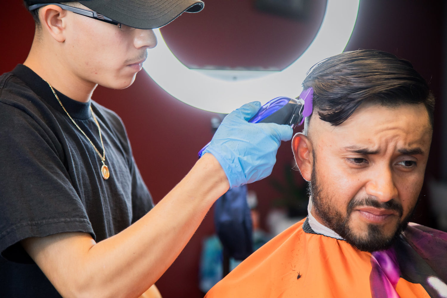 Barber with a black hat and shirt giving a client with an orange apron a haircut