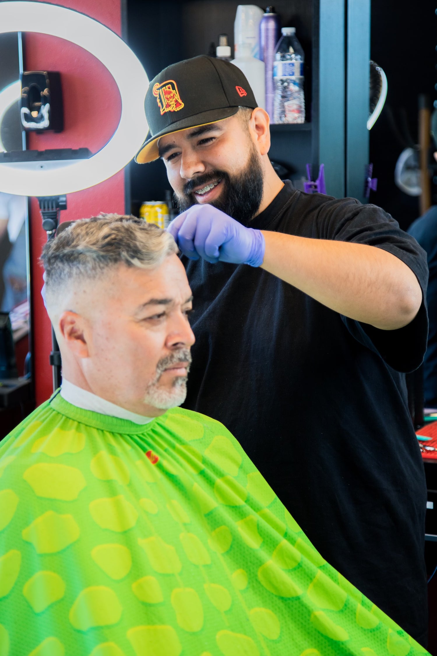 Barber with a black hat and shirt giving a client with a green apron a haircut