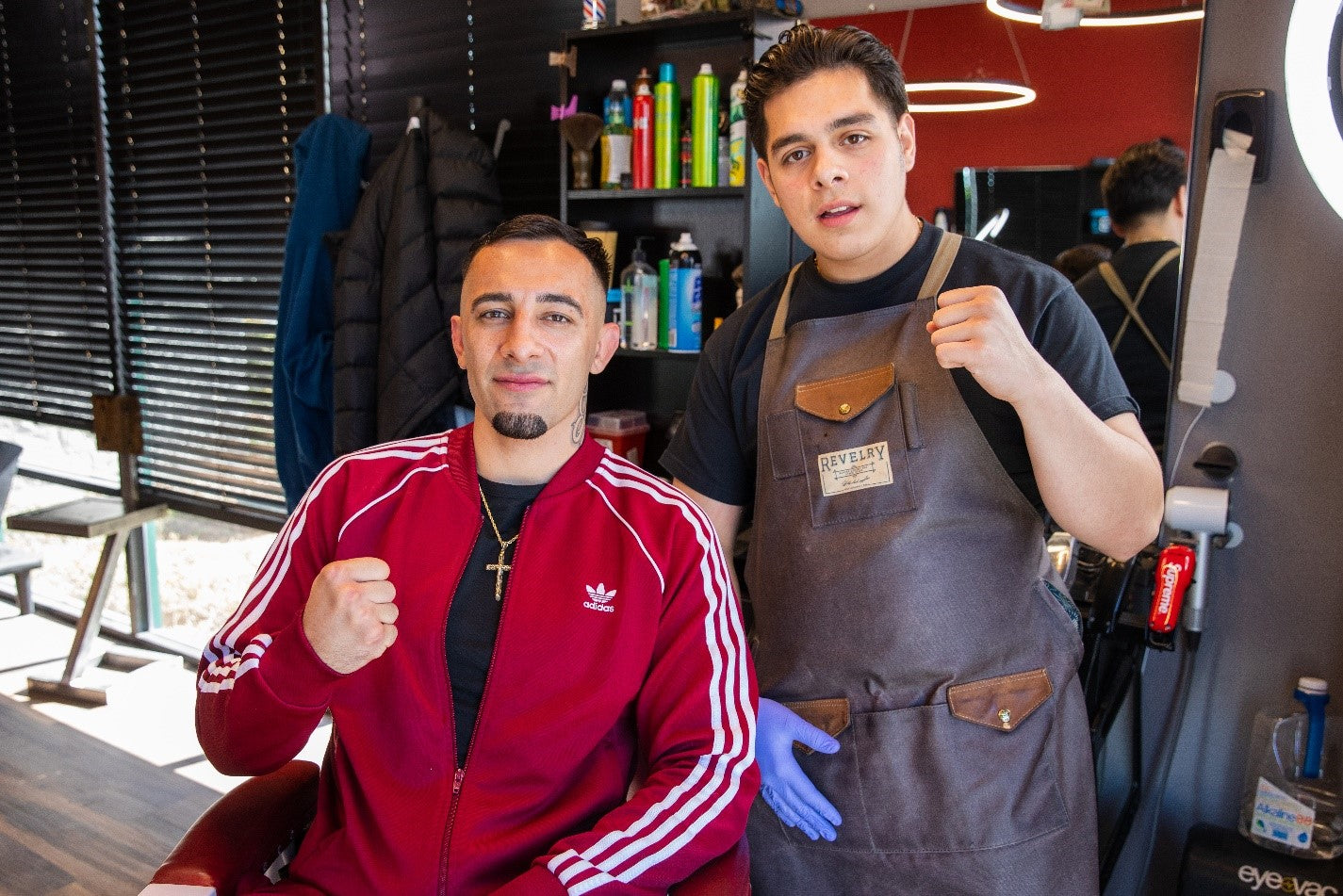 A barber with brown apron standing next to a client in a red sweater