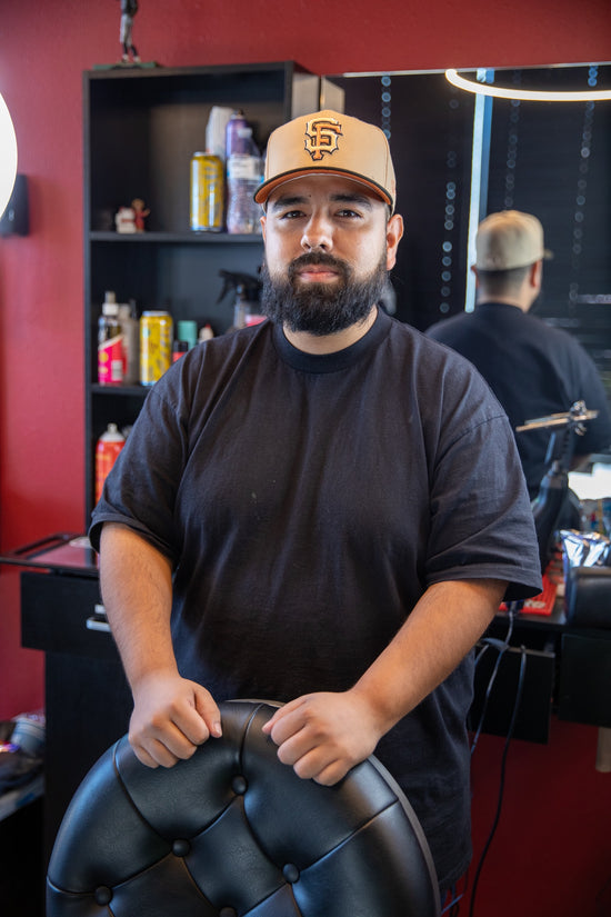 Barber in a black shirt with brown hat leaning on black barber chair